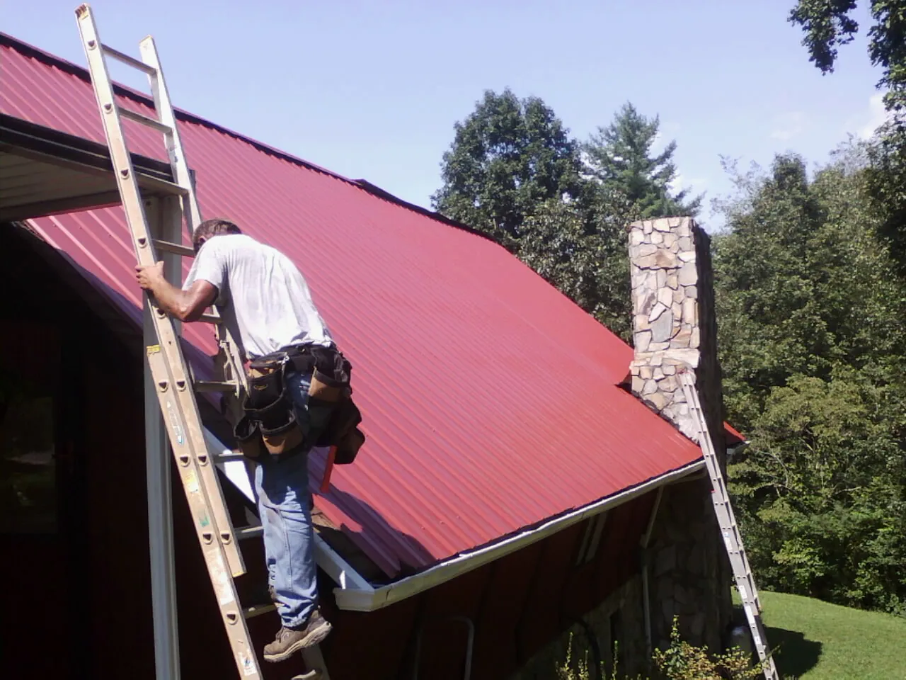 Roofer on a ladder beside a red metal roof and stone chimney — Soffit & Fascia Repair by Roofing Brothers in Palisade